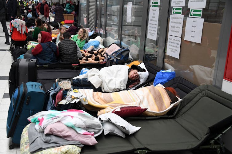 People are sleeping under blankets on beds at a refugee center.