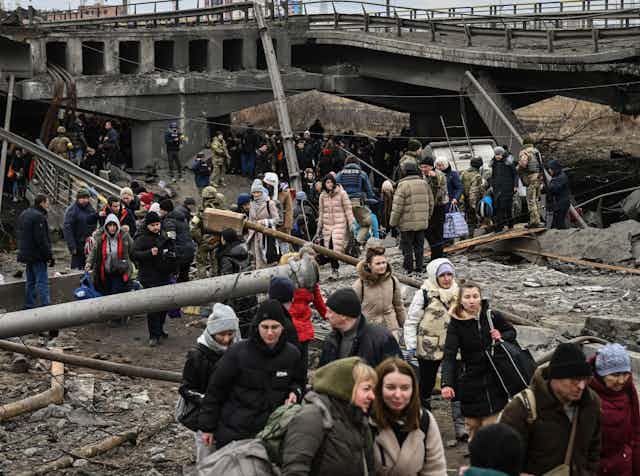 People dressed in winter coats and hats are seen walking pass ruins of a bridge destroyed during Russian invasion. 