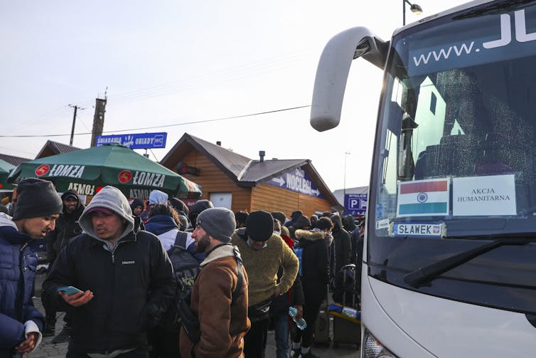 Dozens of men wearing winter coats and hats are shown standing near a bus.