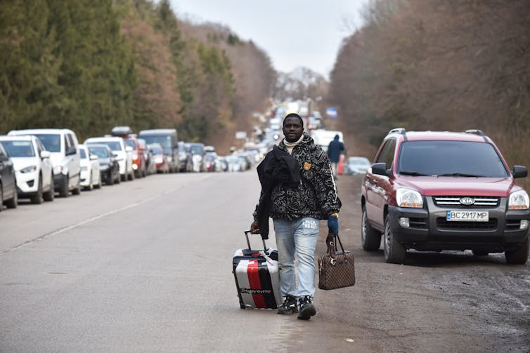 A black man is seen walking with his suitcases along a highway that has two line of parked cars.