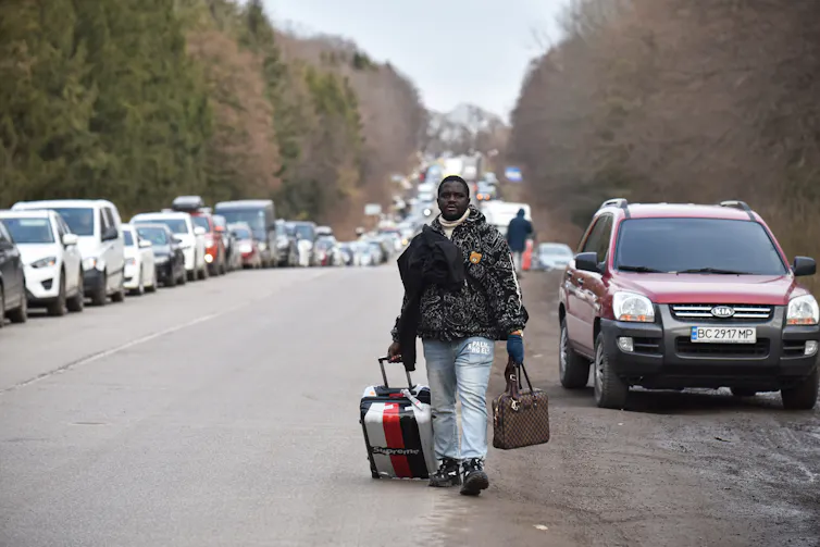 A black man is seen walking with his suitcases along a highway that has two line of parked cars.