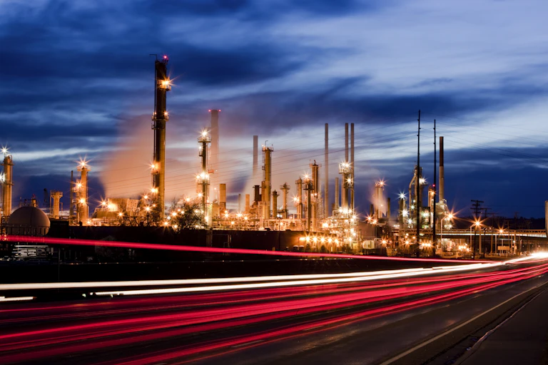 Vehicle taillights pass a refinery light up at night.