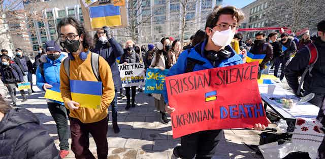 People seen at a protest carrying paper Ukrainian flags and a sign 'Russian silence = Ukrainian death