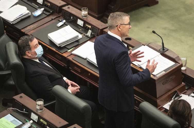 A man stands speaking into a microphone at a podium. Another man sits beside him wearing a blue mask.