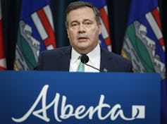 A man speaks into a microphone at a podium with an Alberta banner.