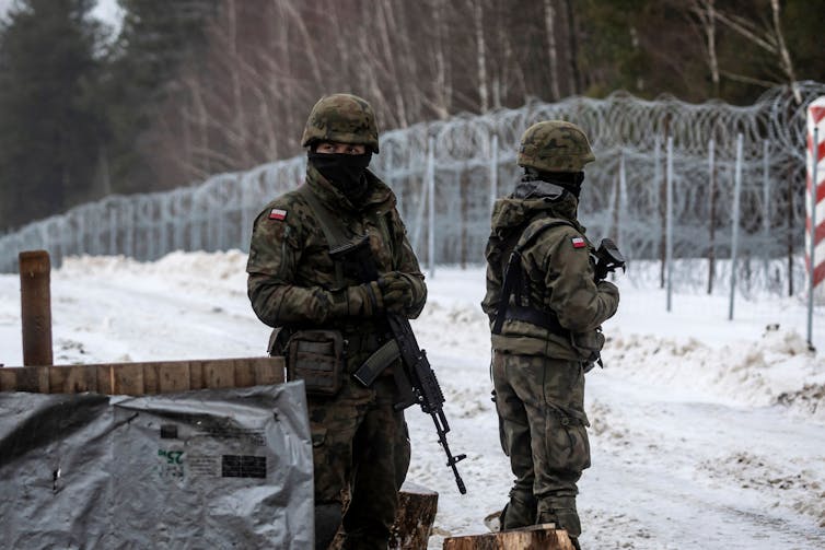 Two soldiers with rifles are guarding a section of woods that is separated by a metal fence with razor-sharp wire attached on top.