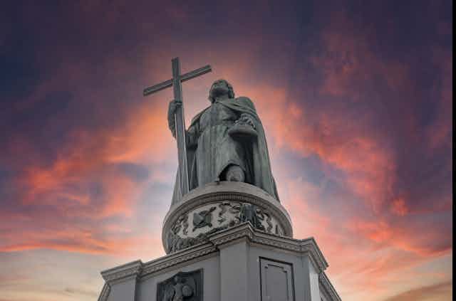 A statue of a man in robes holding a cross is seen against a pink and purple sky.