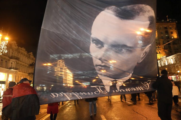 People marching in the streets carry a large banner with a man's face on it.