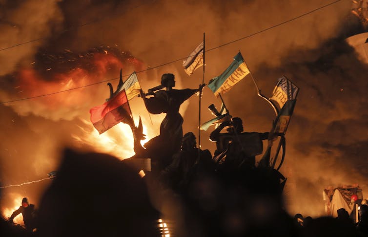 Flames are seen as protesters and riot police clash around a statue silhouetted in the smoke.