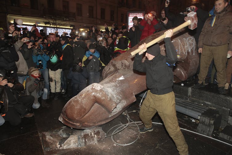 A protester beats a Lenin statue with a sledgehammer as people around him cheer.