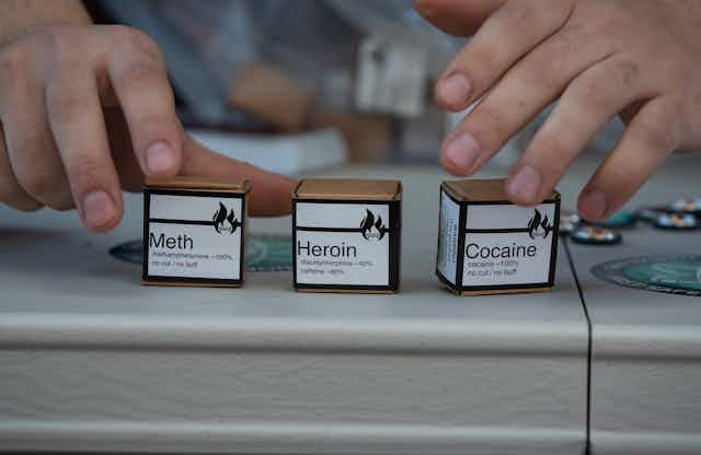 Hands reaching for three small cardboard boxes labeled 'Meth,' 'Heroin' and 'Cocaine.