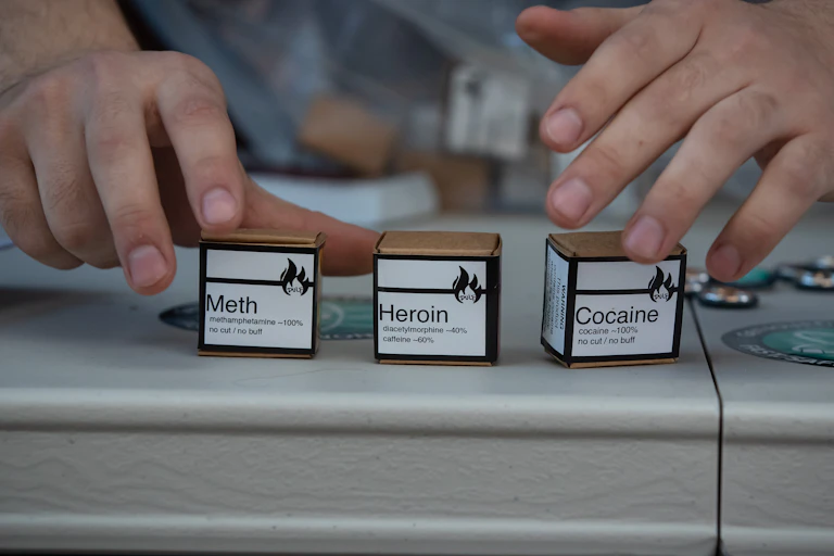 Hands reaching for three small cardboard boxes labeled 'Meth,' 'Heroin' and 'Cocaine.