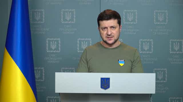 Ukrainian president, Volodymyr Zelensky, recording a video message at a podium, flanked by a Ukrainian flag.