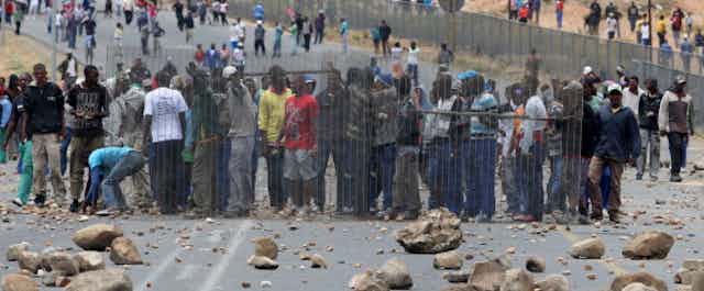 Crowds protesting on a tar road littered with rocks and debris.