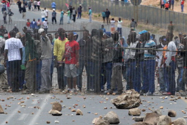 Crowds protesting on a tar road littered with rocks and debris.