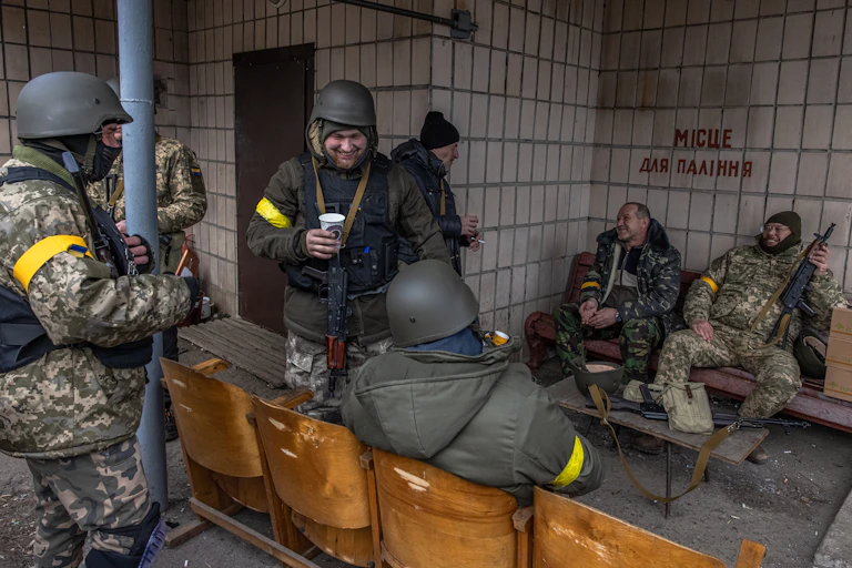 A group of seven men in military uniform and some holding weapons, sitting and socialising at their base