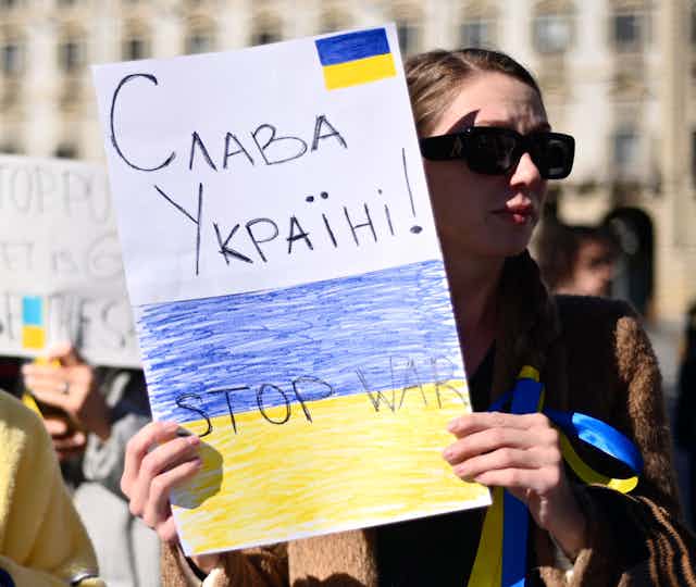 A woman holds a handmade sign that reads слава україні, Stop War and has the Ukrainian flag drawn on it.