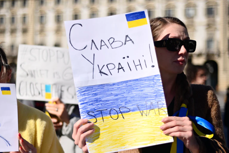 A woman holds a handmade sign that reads слава україні, Stop War and has the Ukrainian flag drawn on it.