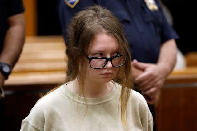 A woman wearing big black glasses and a beige crewneck sits in front of correctional officers.