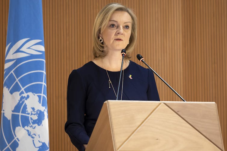 Liz Truss speaks at a podium, with a UN flag behind her.