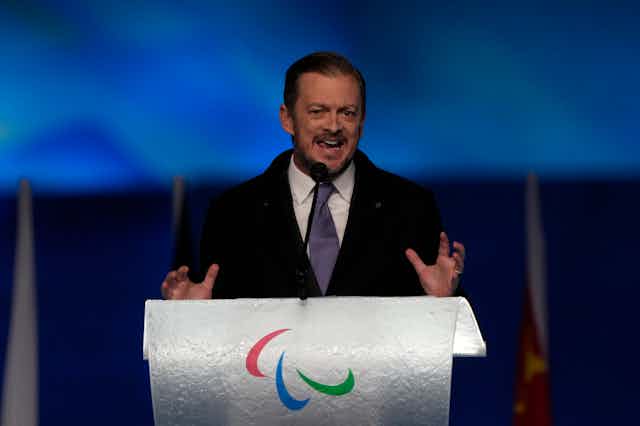 Man in a suit and tie standing behind a podium with Paralympics logo on it
