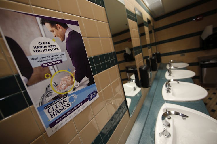 A sign above a row of sinks shows an image of a man washing his hands with the message 'Clean hands keep you healthy. Wash your hands with soap and water for at least 20 seconds. Life is better with clean hands.'