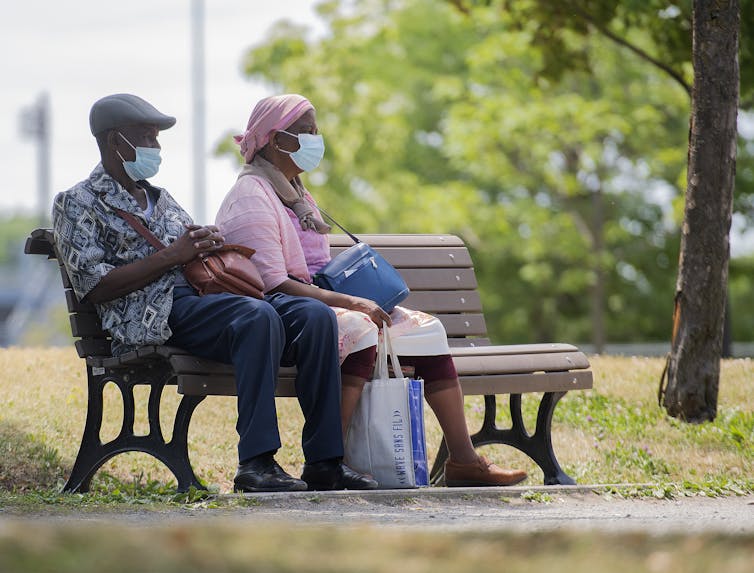 Two people in masks sitting on a park bench.