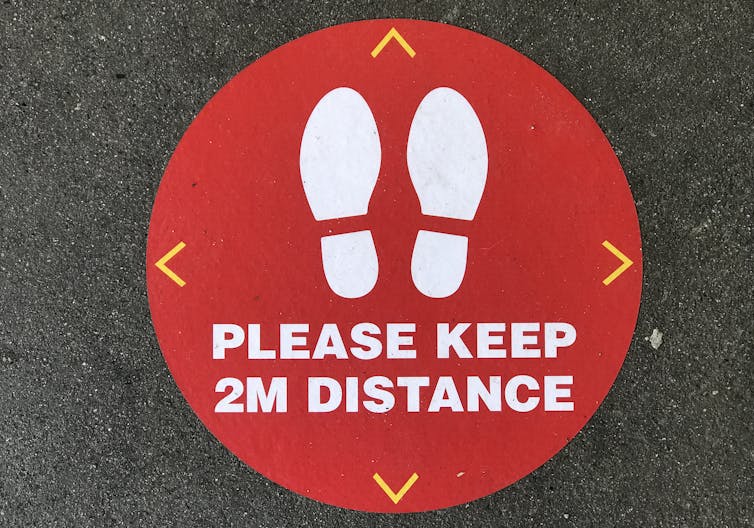 Close-up image of a red social distancing circle on asphalt, with two shoe-prints and the message 'Please keep 2M distance'