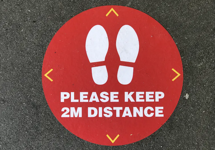 Close-up image of a red social distancing circle on asphalt, with two shoe-prints and the message 'Please keep 2M distance'