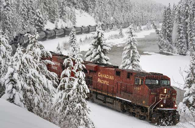 A long train rounds a bend next to a river, surrounded by snow-covered trees.