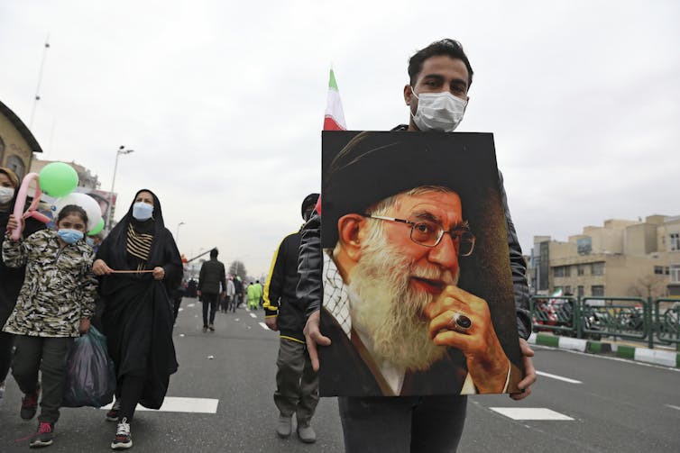 A man wearing a mask carries a poster of the ayatollah during a street march.