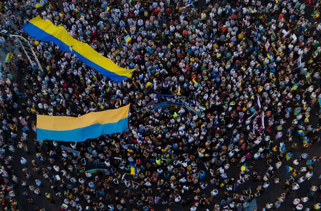 A large crowd of people are seen from above, some carrying two long Ukrainian flags of yellow and blue