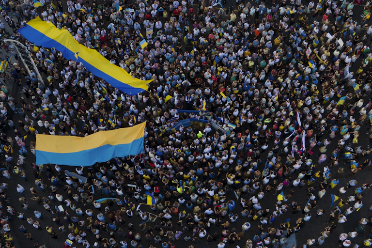 A large crowd of people are seen from above, some carrying two long Ukrainian flags of yellow and blue
