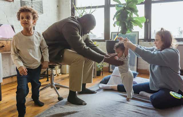 A child seen lunging / stretching while two parents attend to a baby lerarning to stand.