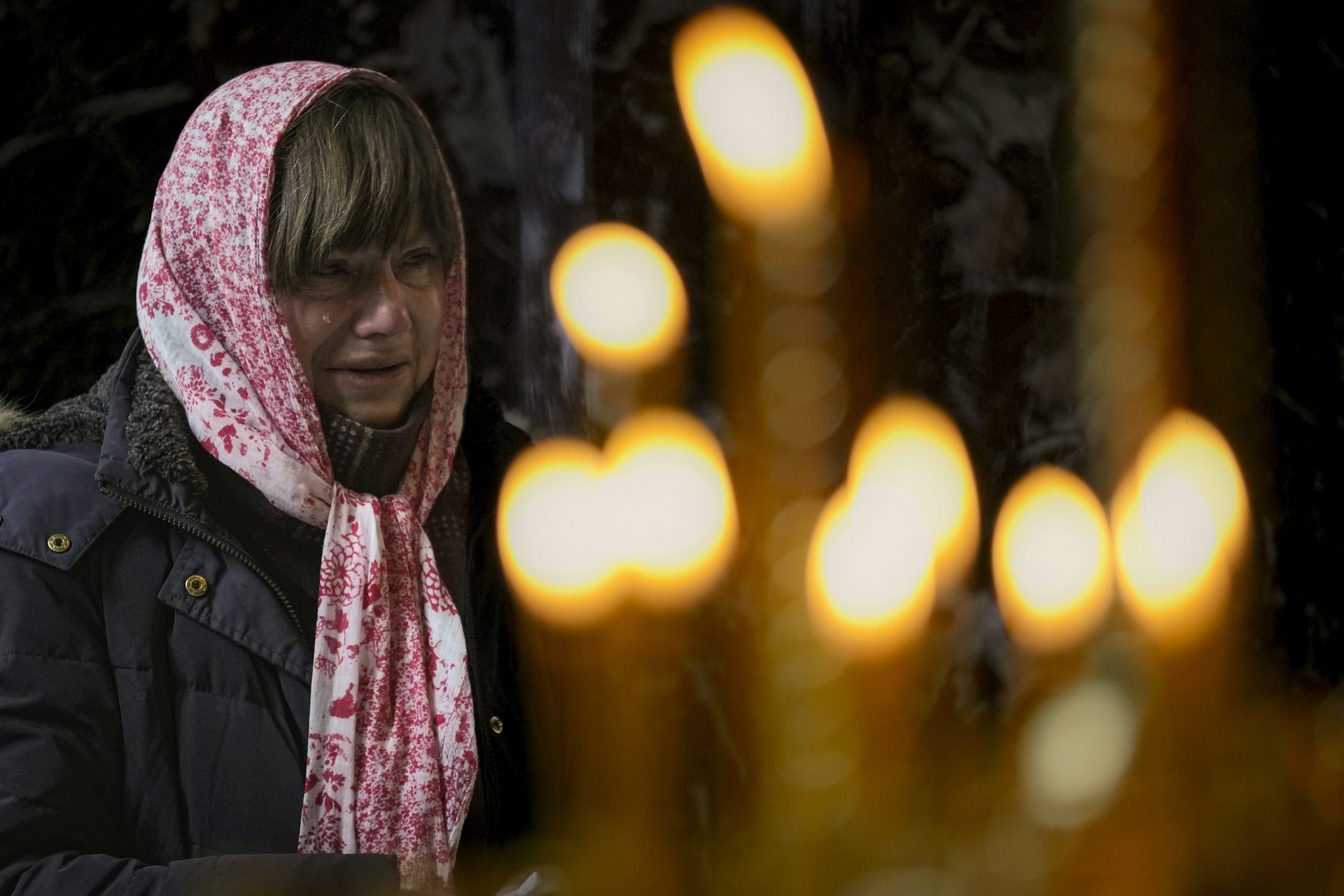 A woman with a flowered scarf on her head weeps in church with candles behind her.