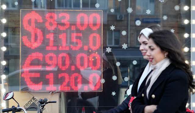 Two women walk in front of a screen displaying the exchange rates of U.S. Dollar and Euro to Russian Rubles