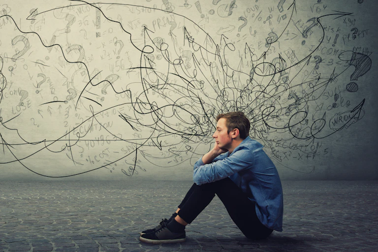 A tired-looking teenage boy sits on the floor with his face resting on one hand, seemingly discouraged, with a wall filled with question marks, exclamation points and scribbles in the background.