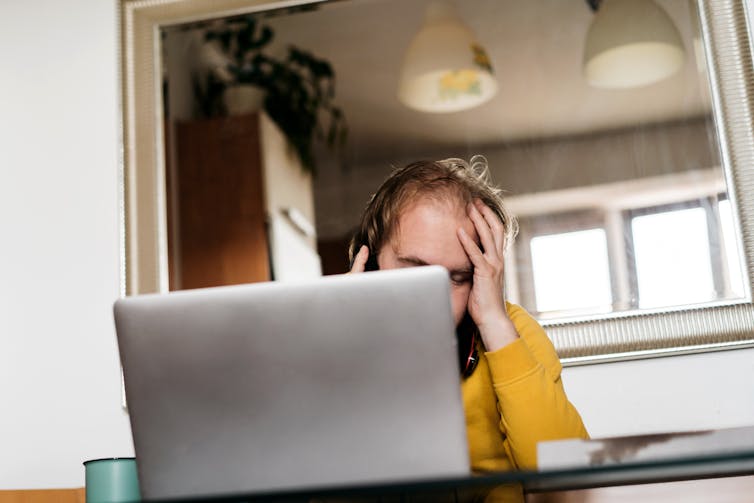 Man at table, face partially obscured by laptop, leaning his forehead into his hand.