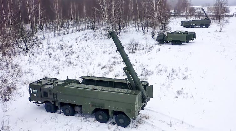 four large military vehicles in a snow-covered field at the edge of a forest, two of the vehicles with nearly vertical cylinders attached at the rear