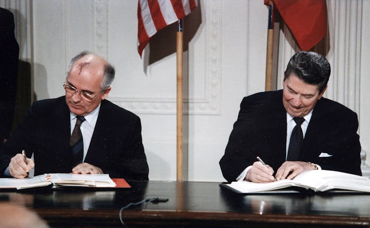 two men in dark business suits sit on the same side of a table signing documents