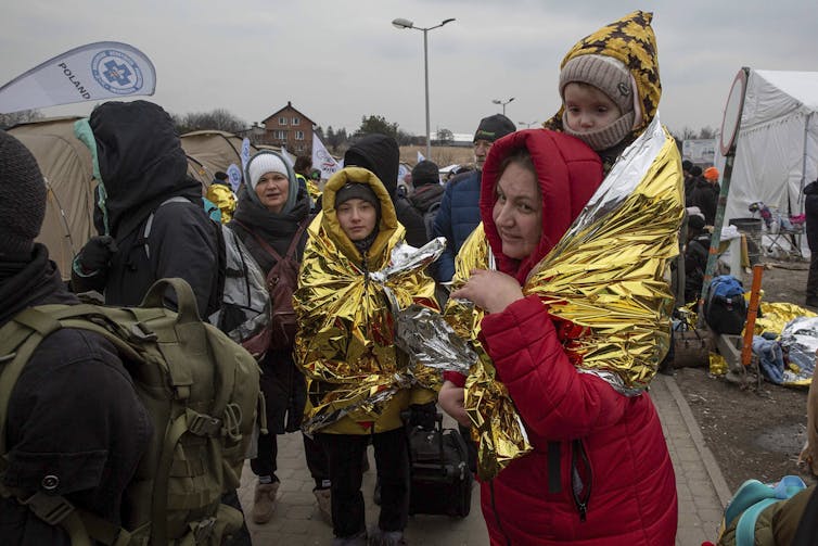 A woman holds a child covered in a thermal blanket on her shoulder in a crowd of other refugees.