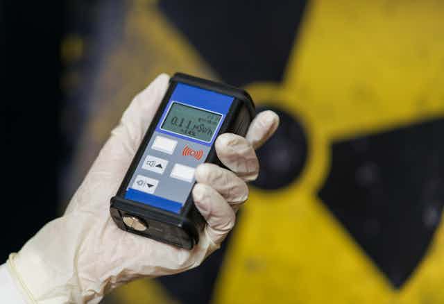 Une main protégée d'un gant tient un compteur Geiger devant le sigle du nucléaire.