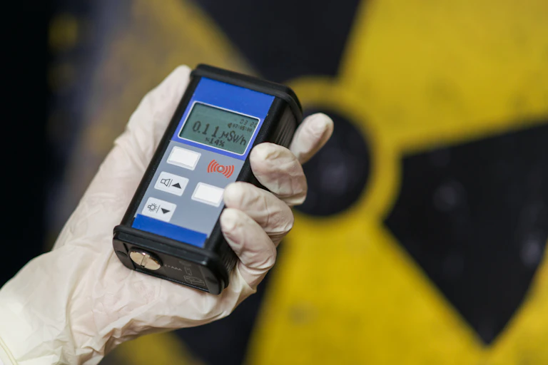 Une main protégée d'un gant tient un compteur Geiger devant le sigle du nucléaire.