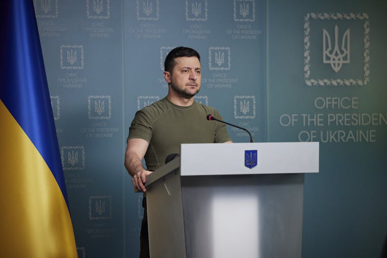 Volodymyr Zelensky in a military green t-shirt standing at a podium with an expression of resolve on his face