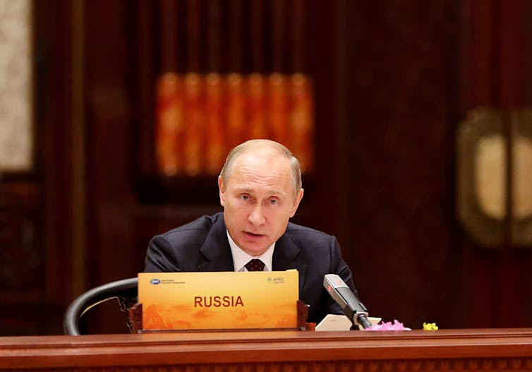 A balding white man in a suit sits behind microphones. In front of him is a sign reading