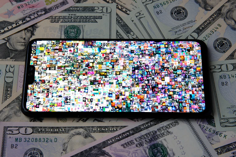 Photo of a mobile phone with Beeple's Everydays artwork on screen, a collage of dozens of tiny photos. The phone is sitting on a pile of US 50 and 20 dollar bills.