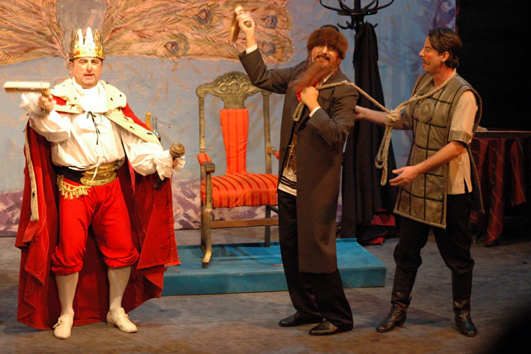 Three actors on stage, with one dressed as a king wearing a crown and a cape, and another dressed as a rabbi, with a long beard.