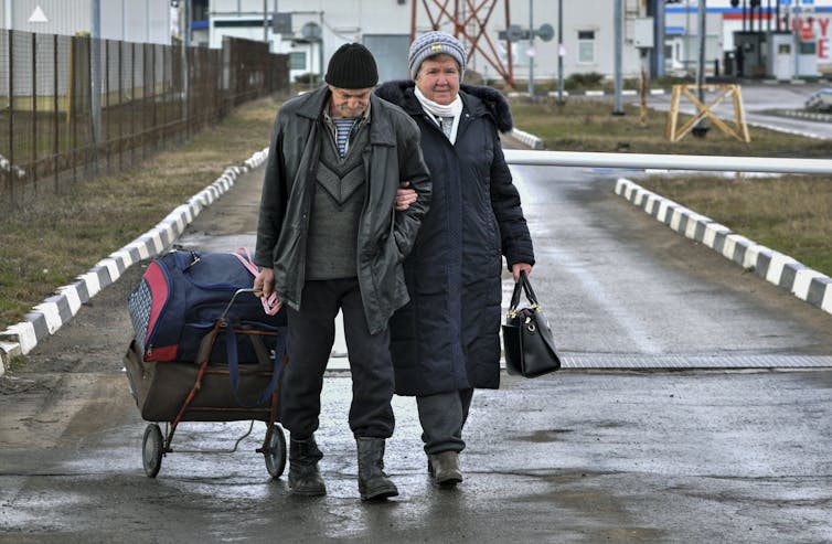 An elderly man and woman with a suitcase walk down road next to wire fence.