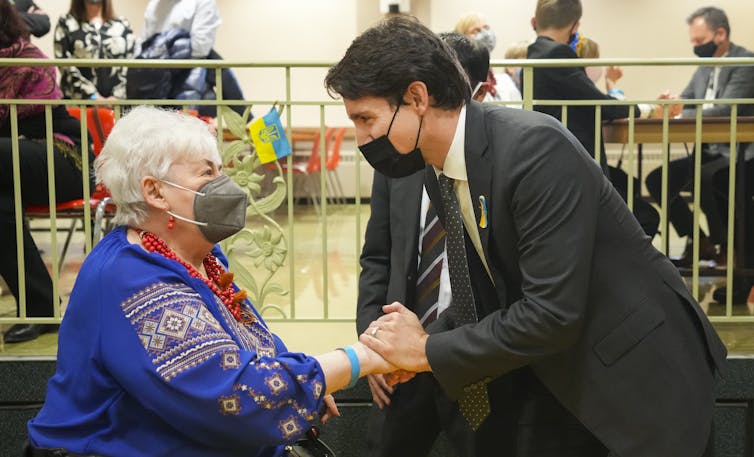 A dark-haired man in a suit wearing a mask shakes holds the hand of a grey-haired woman in a blue patterned sweater.