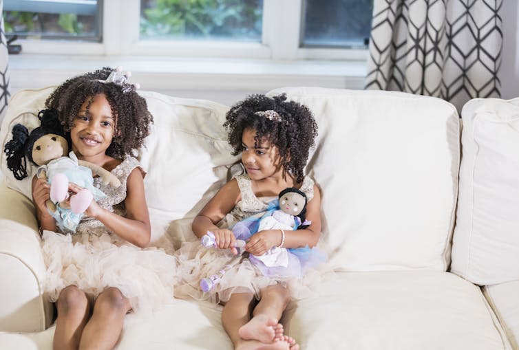 Two Black girls wearing dresses each hold a doll in their hands while sitting on a couch.